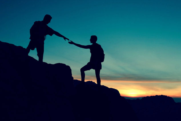 Silhouette of two hikers at sunset, one reaching out to help the other up a rock, symbolizing teamwork or achievement.